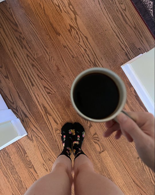 A first-person view looking down at socked feet on a hardwood floor while holding a mug of black coffee, capturing a quiet morning moment that fits the casual "Gm" (good morning) greeting.