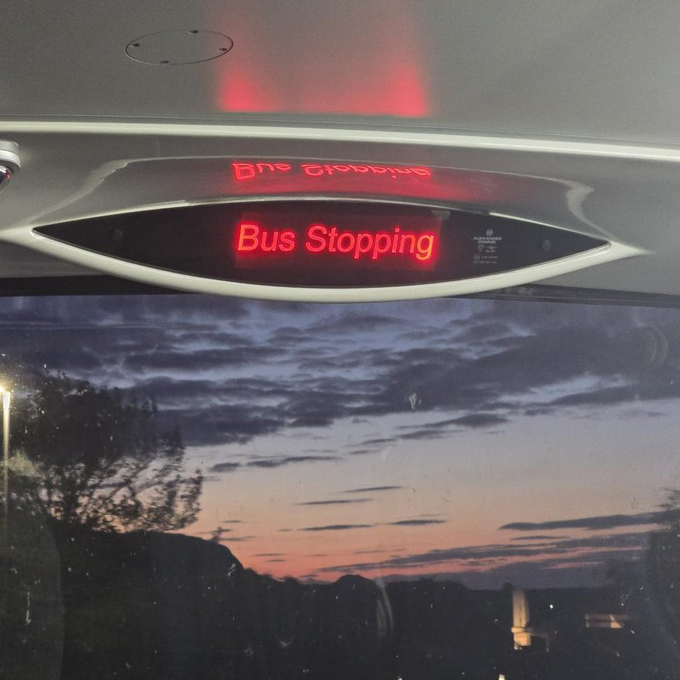 The interior of a bus featuring an LED display sign that humorously reads 'Bus Stopping', with a dramatic sunset sky and silhouetted landscape visible through the window below.