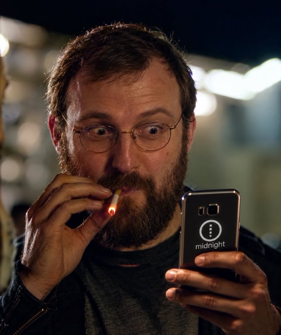 A bearded man with glasses lights a cigarette while looking intently at his phone, which displays a phone case labeled "midnight."

The image captures a candid, dramatic moment of anticipation — his wide-eyed expression and the act of lighting up ("LFG" / "let's go") suggest he's reading exciting news or reacting to something thrilling on his phone, with the cigarette serving as a stress-relief ritual in the moment.