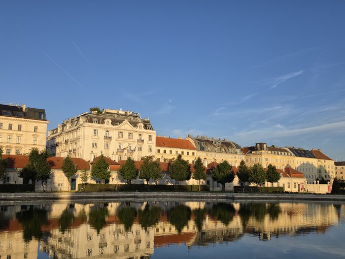 A picturesque waterfront scene shows classic buildings with red-tiled roofs and cream-colored facades mirrored perfectly in the calm water below, set against a clear blue sky with wispy clouds.

The serene view appears to be enjoyed during a leisurely morning abroad, where the traveler is listening to their favorite radio show while enjoying breakfast with a beautiful European cityscape as their backdrop.