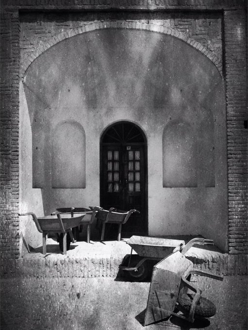 A black and white photograph shows wheelbarrows parked in front of an arched doorway set within a larger brick archway, with a small white number "7" visible above the door. The image captures the quiet, everyday architectural beauty of an ordinary doorway—a fitting subject for the "doors&windows 20" NFT collection by @aghasiani on objkt.com, which appears to celebrate such found doorways and windows as art.