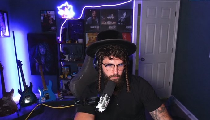 A bearded man with glasses and braided hair wears a black hat and headphones while sitting in a gaming chair in a blue-lit room featuring guitars and framed posters.