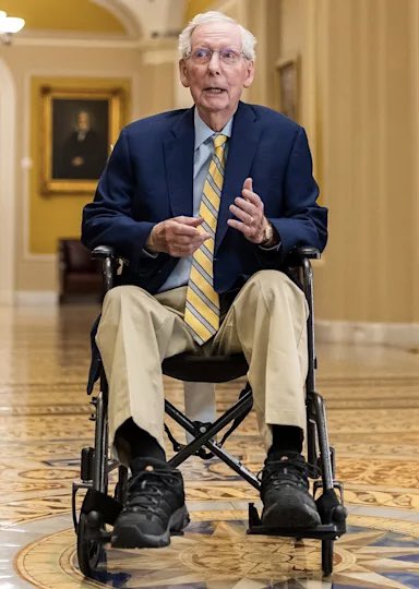 An elderly man in a navy blazer and yellow striped tie sits in a wheelchair in an ornate government building hallway, gesturing while speaking. The post identifies him as 84-year-old Mitch McConnell, who was wheeled out to vote on the Crypto Clarity Act.