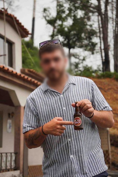 A man with a beard and sunglasses on his head stands outside, pointing at a brown beer bottle labeled 'OKER BOCK' in his other hand.