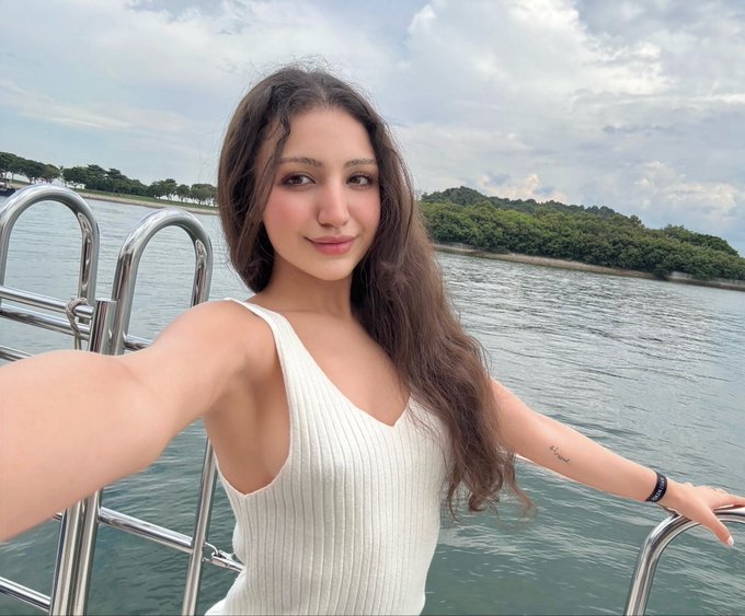 A woman with long dark hair wearing a cream ribbed tank top takes a selfie while leaning against the railing of a boat, with calm water and a tree-lined shore in the background. The serene, sunny setting complements the cheerful "gm (say it back)" greeting, as she beams at the camera with a soft smile in what appears to be a perfect morning on the water.