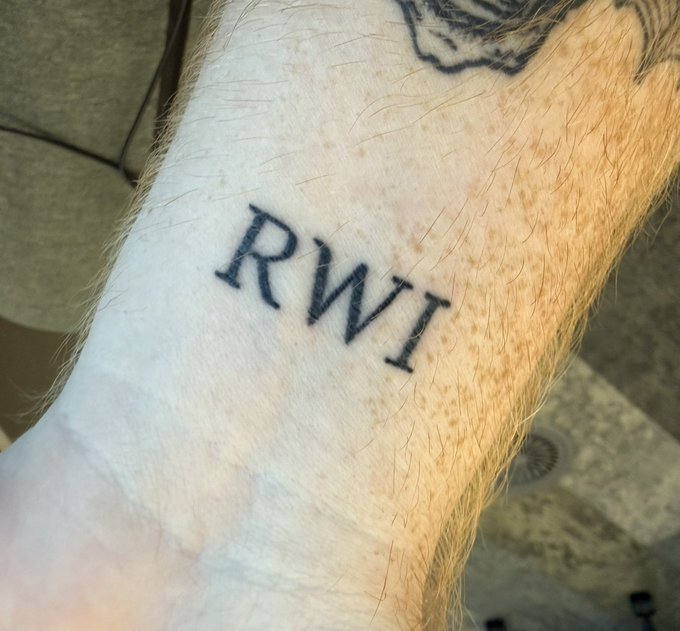 A close-up shows a hairy forearm with the letters "RWI" tattooed in black serif font on the wrist, visually reinforcing the post's claim about the $RWI ticker by humorously comparing skeptics' disbelief in the stock to assuming the tattoo itself is fake.