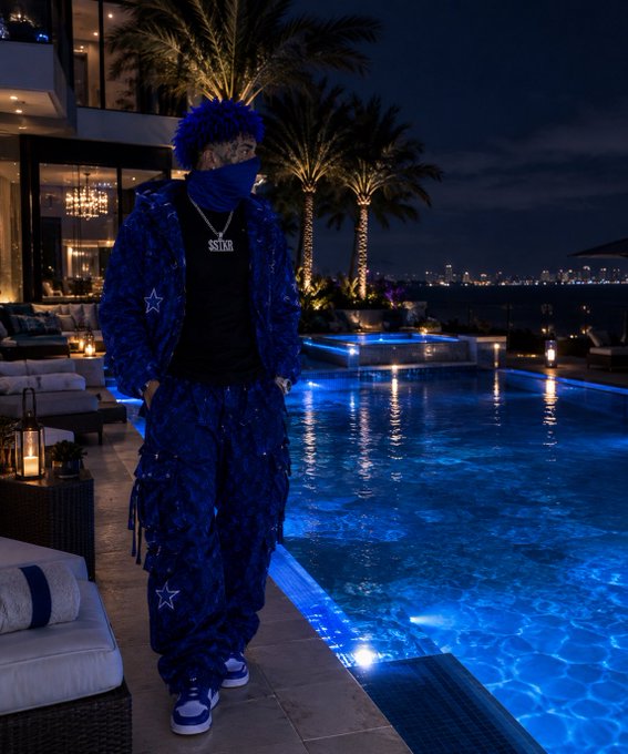 A nighttime photograph of a person wearing a blue camouflage outfit, black t-shirt with 'SOSK' text, and face covering, standing by an illuminated swimming pool with palm trees in the background.