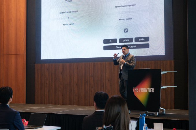 A man in a suit presents on stage at 'THE FRONTIER' conference, standing before a screen showing categories like 'US', 'LATAM', 'EMEA', 'Global', and 'Capital'.
