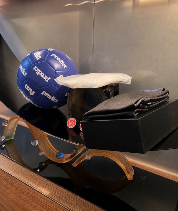 A display shelf showing a blue 'Predict' soccer ball, orange sunglasses, a black wallet, and other branded merchandise on a reflective surface.