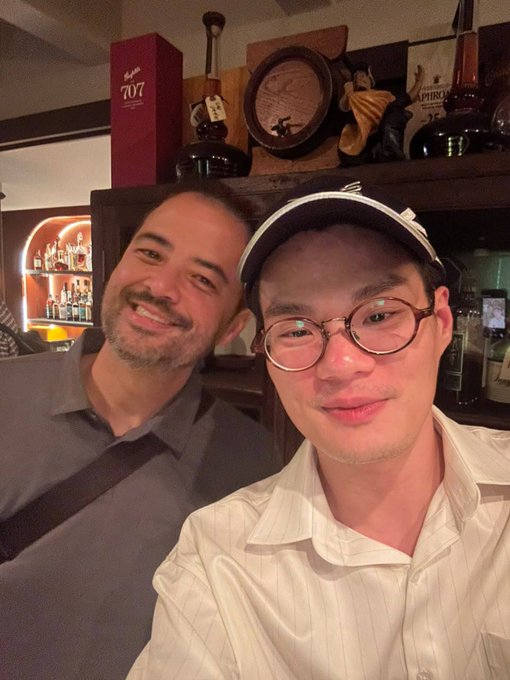Two friends pose for a selfie at what appears to be a bar or lounge, with the man on the right wearing a white cap, round tortoiseshell glasses, and a striped button-up shirt, while the man on the left smiles in a gray polo shirt. Behind them, a shelf displays liquor bottles and a red box labeled "707," fitting the caption's vibe of a fun night out in Singapore during Token 2049.