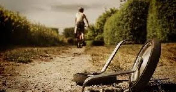 A sepia-toned photograph of a person riding a bicycle away down a dirt path, with an overturned wheelchair lying in the foreground.