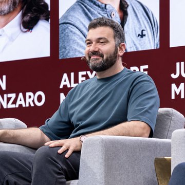 A smiling man with a beard sits on stage at the #SuiBasecamp2026 conference, with event posters featuring names like "ALEX" and "NZARO" visible behind him, relating to the post about Alexandre Moufarek from GoogleDeepMind speaking at the event.