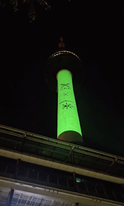 A tall observation tower is illuminated in bright green at night, featuring projected logos including "Zeit" and the Wikipedia emblem on its shaft. The structure appears to be Kyoto Tower, viewed from below against the dark sky.