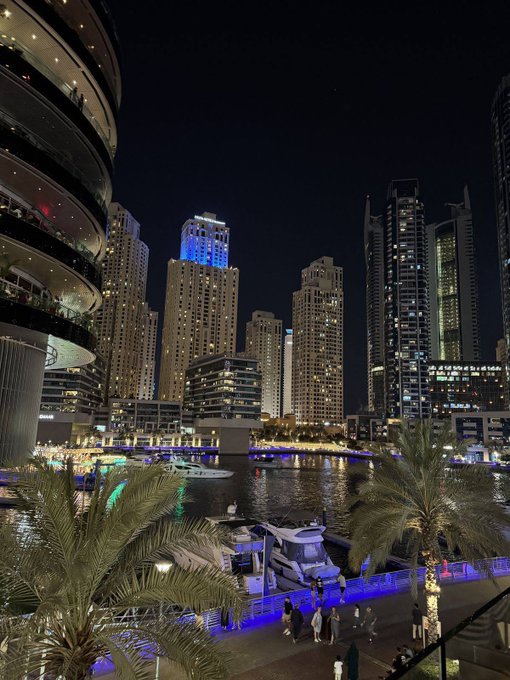 A nighttime view of Dubai Marina showing illuminated skyscrapers, boats docked in the harbor, and a building with a glowing curved facade on the left.