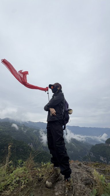 A hiker wearing dark clothing and a mask stands on a rocky mountain summit with a flowing red scarf/sash in the wind, overlooking misty mountains and clouds.