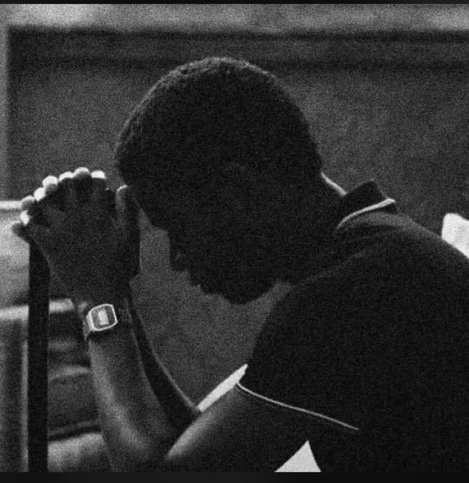 A black and white image shows a man in a dark polo shirt with his head bowed and hands clasped together in prayer or contemplation, wearing a digital watch. The somber, reflective pose contrasts with the post's upbeat "let's get that bag" message, suggesting a moment of quiet preparation or gratitude before the work week begins.