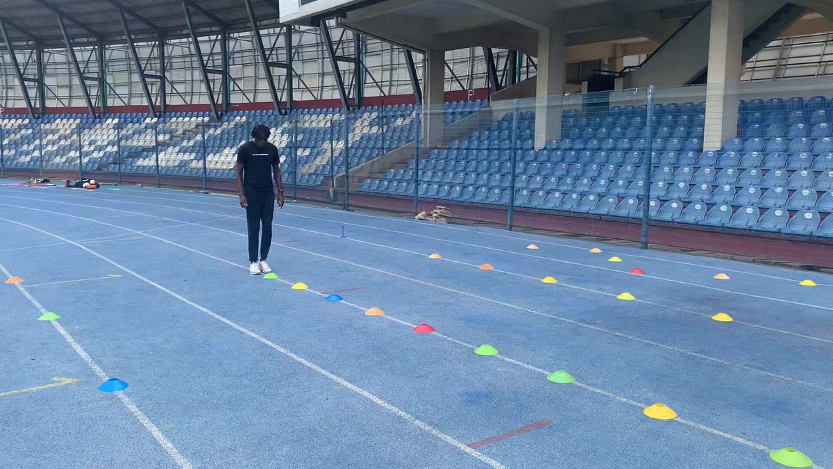 The image shows a man standing on an indoor track, wearing black clothing and holding a stop sign. He appears to be instructing or demonstrating some form of exercise technique to others who are present at the event. The setting seems to be indoors with blue bleachers in the background.