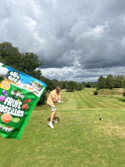 A golfer in an orange shirt and shorts swings a club on a grassy course under a cloudy sky, with a large package of Rowntree's Fruit Pastilles candy edited into the foreground.