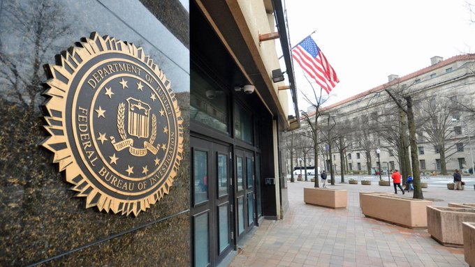 The seal of the Department of Justice Federal Bureau of Investigation (FBI) is mounted on a marble wall outside a building with an American flag, where pedestrians walk on a city sidewalk.