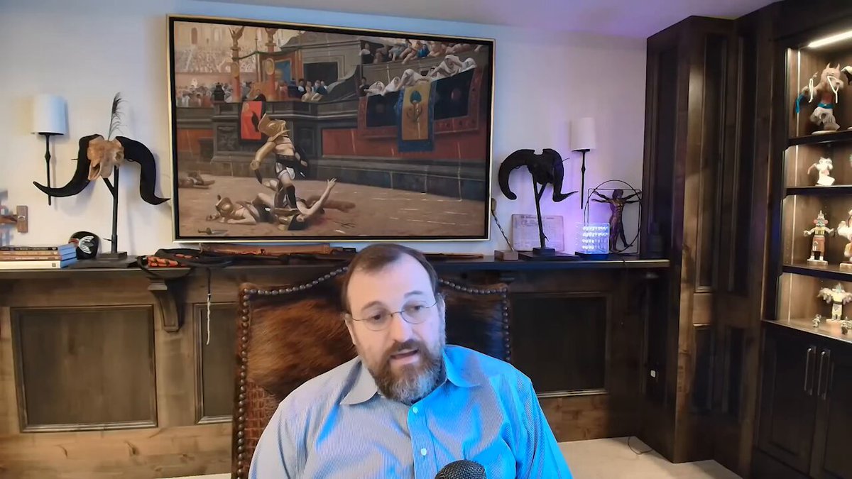 The image shows a man sitting in an office chair with his legs crossed while wearing glasses. He appears to be talking or giving a presentation, as indicated by him looking at the camera. The room has a desk and shelves filled with various items such as books and figurines. There is also a painting on the wall above the man's head.