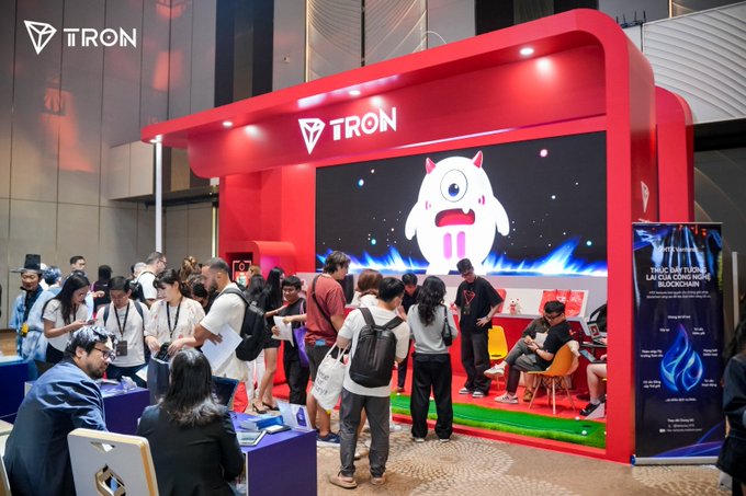 A crowded TRON booth at an industry event features the company's logo and a mascot displayed on a large screen as attendees network and engage with staff.