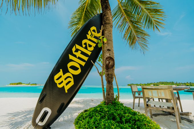 A "$OLHARE" surfboard leans against a palm tree on a tropical beach, perfectly capturing the sunny, laid-back vibe of "Solflare Szn." The wordplay between "Solflare" and "surf" pairs the crypto ticker with the iconic beach scene.
