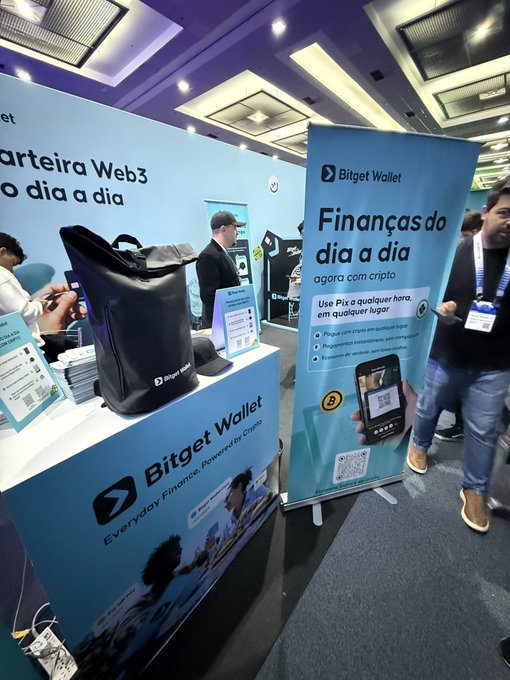 A trade show booth promotes 'Bitget Wallet' with Portuguese text reading 'Carteira Web3' and 'Finanças do dia a dia agora com cripto', featuring a display of branded merchandise.