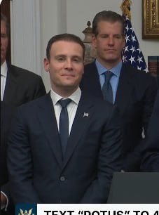 A photograph of a man in a dark suit and tie standing at a podium, with other officials and an American flag visible in the background, with text overlay reading 'TEXT 'POTUS' to 4'.