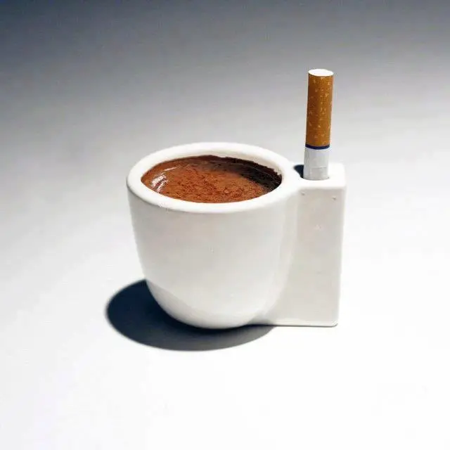 A ceramic novelty ashtray designed to resemble a white coffee mug filled with brewed coffee, featuring a built-in holder on the side that cradles a lit cigarette.
