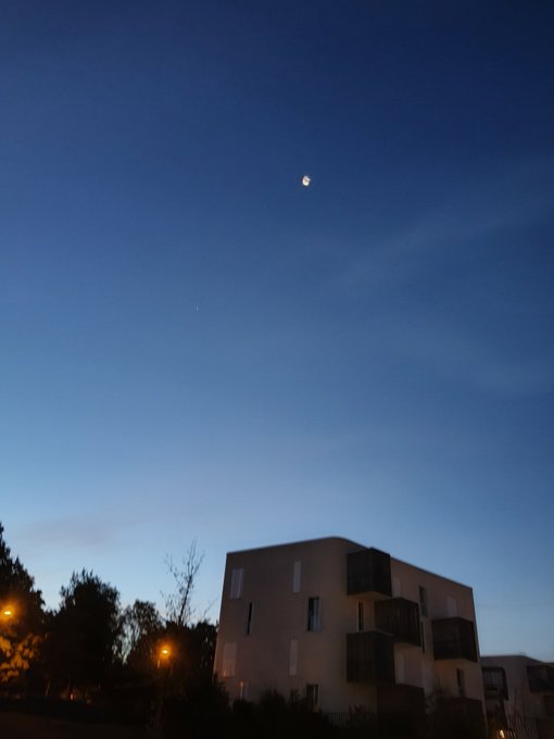 A twilight sky shows the moon and what appears to be a star or planet above a modern building and silhouetted trees.