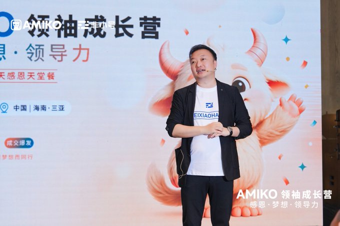 A man wearing a headset microphone and a white 'EIXIAOHA' t-shirt speaks on stage at an 'AMIKO 领袖成长营' event in 中国·海南·三亚, with a cartoon illustration of a horned animal behind him.