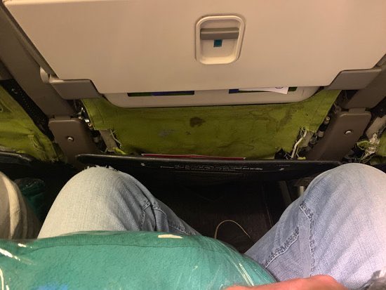 A passenger sits in a cramped airplane seat, their jeans-clad knees pressed close together, with the exposed yellow-green insulation of the seat back in front revealing the aircraft's interior padding. The view captures the unglamorous, tight quarters of economy class travel—fitting given the poster's boastful "blessed arrival in Singapore" and promises to "shill garbage coins," suggesting the trip itself may feel less glamorous than the bragging implies.