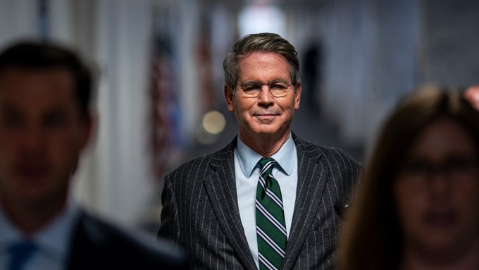 A man in a pinstripe suit with a green striped tie and glasses stands in what appears to be a government hallway with American flags in the background, flanked by two blurred figures in the foreground.