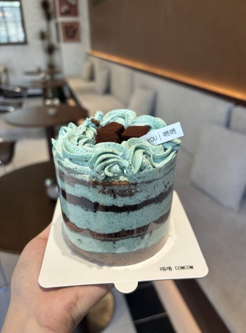 The image features a hand holding up a small cake with blue frosting, placed on a white plate. The cake has chocolate layers and appears to be decorated with icing flowers. The background of the image shows a dining table surrounded by chairs, suggesting that this scene takes place in a restaurant or similar setting. There is also an additional text present in the image, which reads "Crypto Survival Plan | Day 8懒得做饭", indicating that it might be related to cryptocurrency or financial planning.