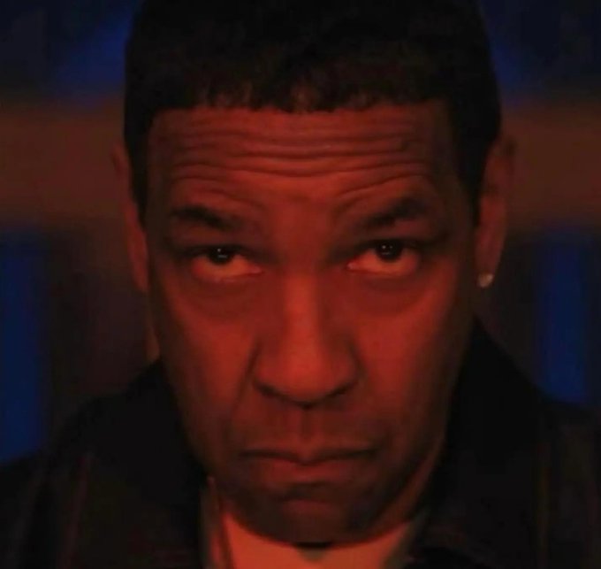 A close-up still of a man with short dark hair staring intensely at the camera under red lighting.