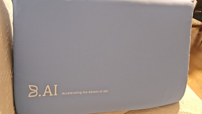 A close-up of a light blue laptop or device lid displaying the 'B.AI' logo and the tagline 'Accelerating the Advent of AGI'.