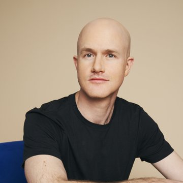 A bald man in a black t-shirt looks directly at the camera against a plain beige background.