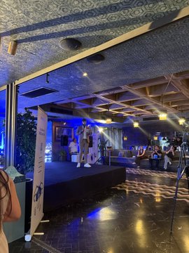 The image shows a large room with a stage at its center, where people are gathered for an event. There are several individuals standing around the stage, some of them wearing ties. The room has a blue ceiling that adds color to the scene. A person is holding a camera in their hand, possibly capturing memories from this gathering or documenting the event.