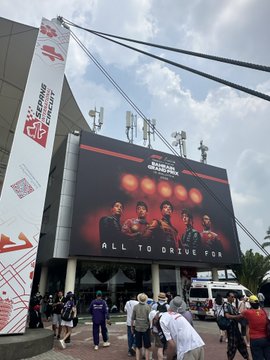 An outdoor scene shows a large digital billboard advertising an esports tournament with players, reading 'ALL TO DRIVE FOR', as people walk by on a sunny day.