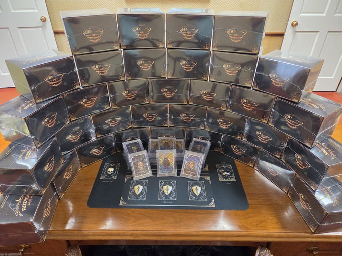 A pyramid of black "$FUZZY" card boxes with gold crests sits on a wooden table, surrounding a display of trading cards featuring character artwork and a playmat with the "Unleash Your Force" slogan, tying into the @fuzzy_xrp trading card promotion.