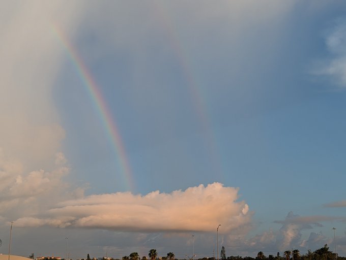 A double rainbow arcs across a partly cloudy blue sky above a large cumulus cloud, with a flat landscape and streetlights visible at the bottom of the frame.