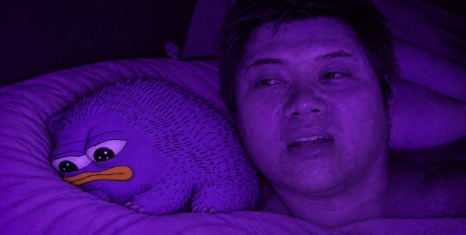 A person lying in bed under purple lighting next to a plush duck toy.
