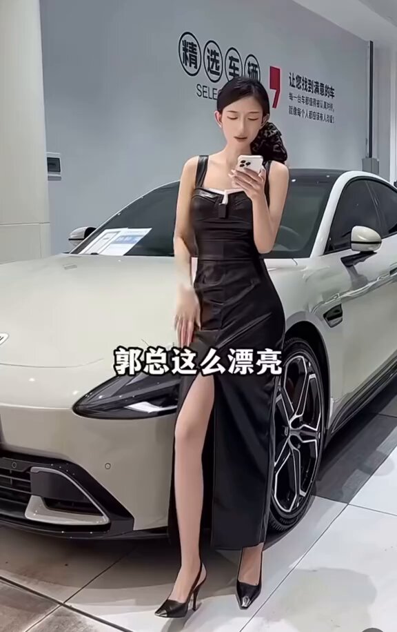 The image features a woman posing next to a car, possibly at an auto show or dealership. She is wearing a black dress with a high slit and heels, giving her a stylish appearance. The car she's standing beside has a large rear tire and a smaller front tire, which are both visible in the photo. The background of the image includes a sign that reads "6.25梭哈晨报", possibly indicating an event or location related to the car show or dealership where the woman is posing with the vehicle.