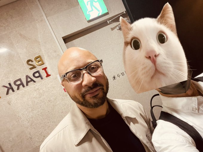A bald man with glasses poses for a selfie next to someone wearing a cat-face mask, with 'IPARK 2F' visible on the wall behind them.