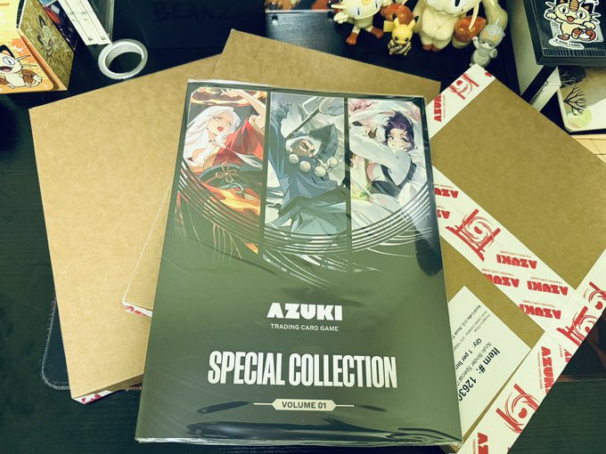 An AZUKI Trading Card Game 'Special Collection Volume 01' box resting on cardboard shipping packages, with Pokémon plush figures visible in the background.