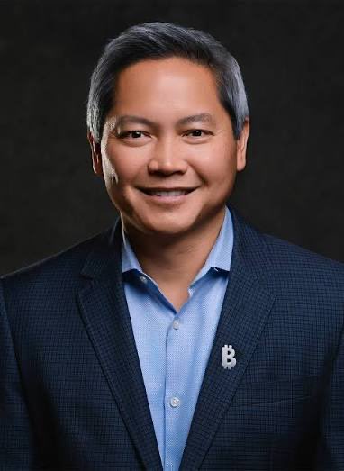 A professional headshot of a man with dark hair wearing a blue shirt and dark blazer, smiling against a black background, with a small Bitcoin "₿" logo pin on his lapel. The Bitcoin-themed attire and confident pose align with the post's bullish $MSTR (MicroStrategy) message highlighting two top crypto-related investments.