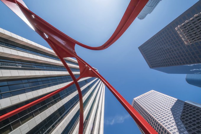 A dramatic upward-angle photograph of modern skyscrapers against a blue sky, framed by a striking red sculptural ribbon weaving between the buildings.