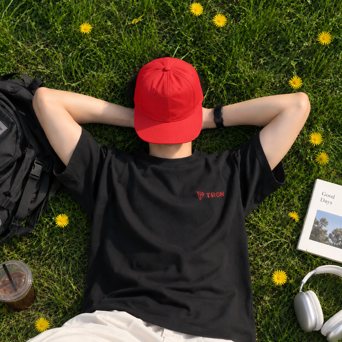 An overhead view of a person lying in grass among dandelions, wearing a red cap and a black t-shirt branded with a red 'TRON' logo, with a backpack, iced coffee, a 'Good Days' book, and white headphones nearby.