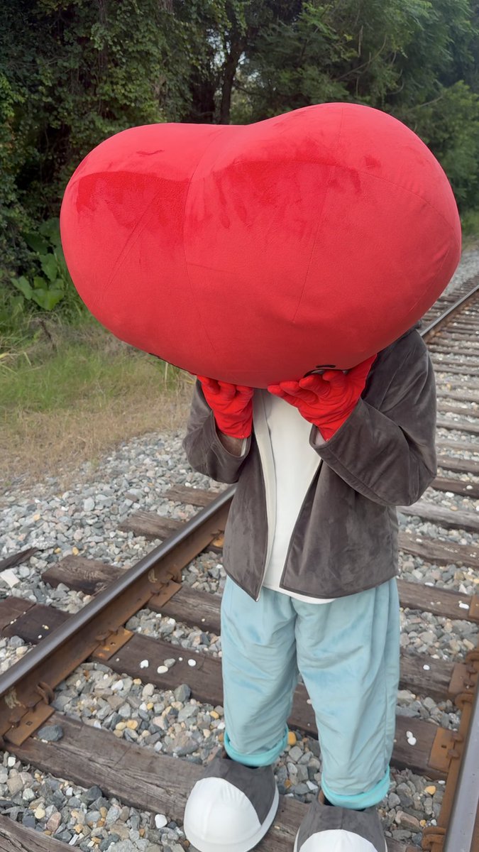 A surreal photograph shows a person standing on railroad tracks with an oversized red heart obscuring their head, wearing red gloves, a brown jacket, and blue pants.