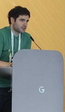 A young man wearing a green shirt stands behind a podium marked with a large "G" (Google logo), speaking into a microphone during what appears to be a presentation at Google, connecting to the post about IOG members visiting Google to discuss Cardano.